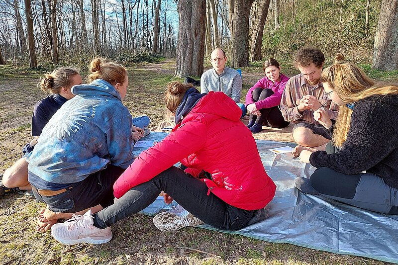 Eien Gruppe von jungen Menschen sitzt auf einer Decke in der Sonne, sie unterhalten sich miteinander.