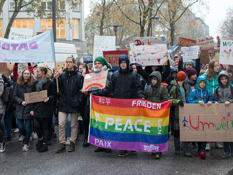 Zahlreiche junge Menschen am Anfang eines Demonstrationszuges. Sie tragen Plakate und Banner mit Aufschriften wie beispielsweise: Peace, Busstag, Umweltschutz.