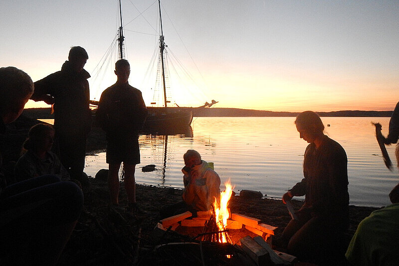 Im Vordergrund mehrere Silhouetten von Personen um ein Lagerfeuer, im Hintergrund ein Segelschiff auf dem Wasser im Sonnenuntergang.