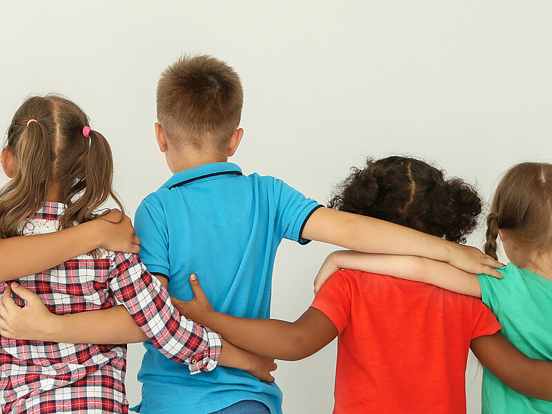 4 Kinder, verschiedener Hautfarbe, in bunter Kleidung, stehen vor einem grauen Hintergrund. Zusehen sind sie von hinten. Sie stehen nebeneinander und haben einander die Arme auf die Schultern gelegt.