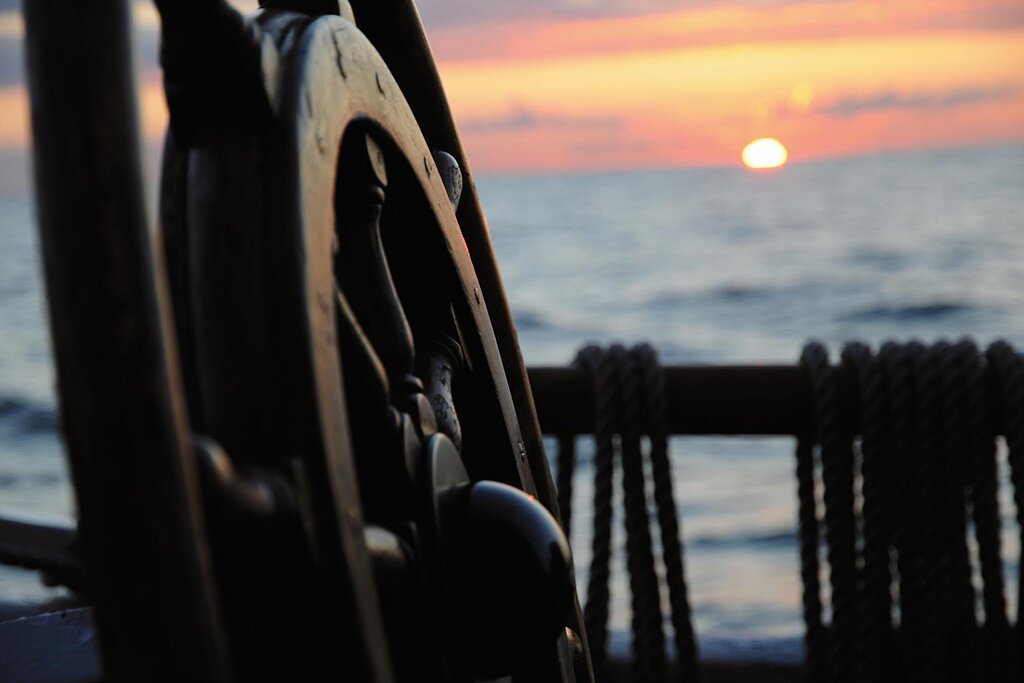 Im Vordergrund ein hölzernes Steuerrad eines Segelschiffs, im Hintergrund ein Sonnenuntergang auf dem Meer.